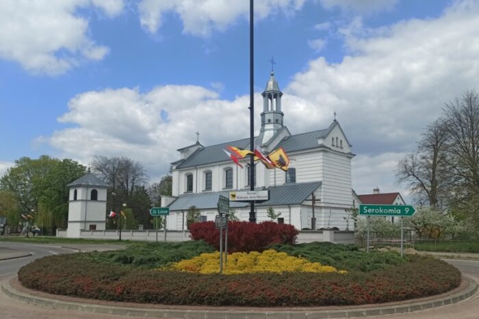 Kościół pod wezwaniem Podwyższenia Krzyża Świętego w Adamowie, na pierwszym planie rondo biskupa Jana Mazura, na słupie powiewają flagi narodowe i gminne, w głębi po lewej dzwonnica